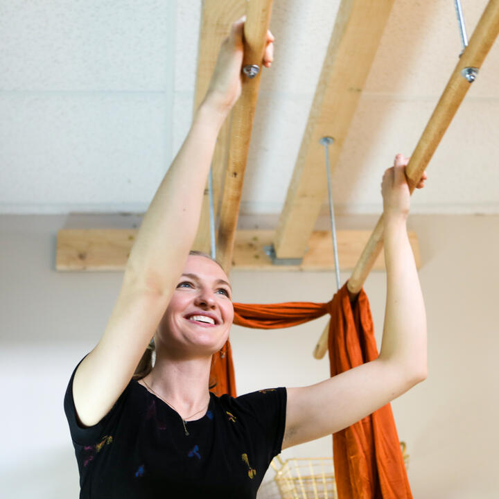 Headshot of the massage therapist, Jordan, holding the parallel Ashiatsu bars that hang from wooden beams over her head.